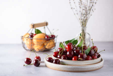 Fresh organic cherries in a gray ceramic plate on a light gray background. In the background there is a basket with apricots and cherries and a saucer with cherries.の写真素材