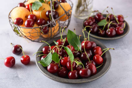 Fresh organic cherries in a gray ceramic plate on a light gray background. In the background there is a basket with apricots and cherries and a saucer with cherries. upper angle.の写真素材