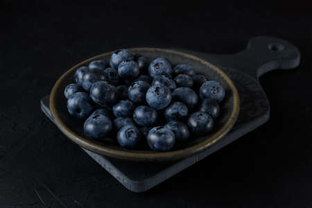Fresh organic blueberries in a ceramic plate. The plate stands on a gray board on a black background. close-up.の写真素材