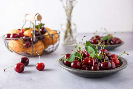 A fresh ripe cherry with leaves lies in a gray ceramic plate. In the background there is a basket with apricots and cherries and a saucer with cherries. close-up.の写真素材