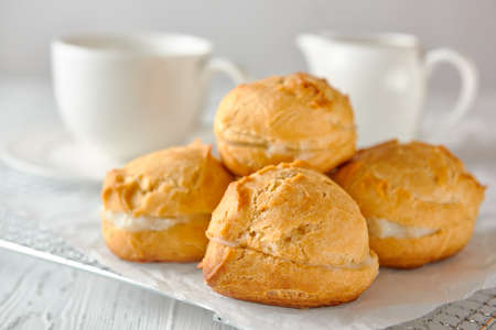 Delicious homemade profiteroles or eclairs on a metal grid covered with white paper. In the background is a teacup and a milk jug. close-up.の写真素材