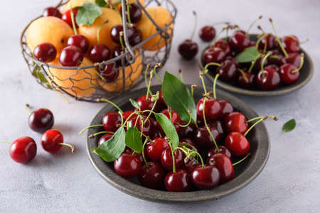 Fresh red cherry with green leaves in a gray ceramic plate. In the background, cherries and apricots in an iron basket and cherries in a ceramic saucer. close-up.の写真素材