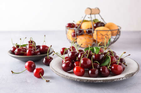 Fresh ripe red cherries in a white ceramic plate. In the background, cherries and apricots in an iron basket and cherries in a ceramic saucer. close-up.の写真素材
