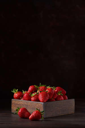 Fresh delicious strawberries in a square wooden bowl on a brown wooden table. Brown background, vertical. Close up, copy space.の写真素材