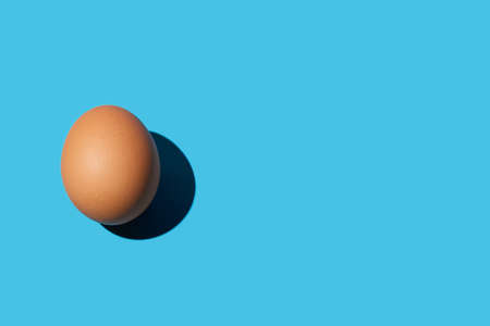 A brown chicken egg lies on the left on a bright blue background. Bright light, deep shadow. Close up, copy space, top view.の写真素材