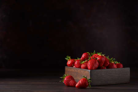 Fresh red ripe strawberries in a square wooden bowl on a brown wooden table. There are two strawberries nearby. Brown background, horizontal. Close up, copy space.の写真素材