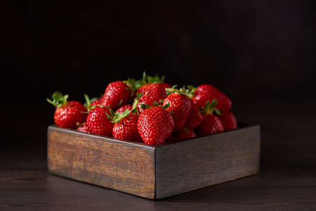 A square wooden bowl filled with fresh strawberries stands on a brown wooden table. Brown background, horizontal. Close up, copy space.の写真素材