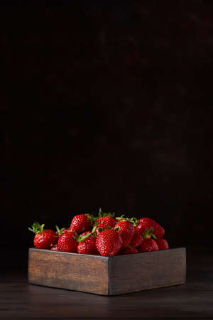 Fresh red strawberries in a square wooden bowl standing on a brown wooden table. Brown background, vertical. Close up, copy space.の写真素材