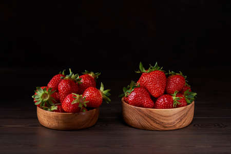 Ripe strawberries in two wooden bowls standing on a brown wooden table. Brown background, vertical. Close up, copy space.の写真素材