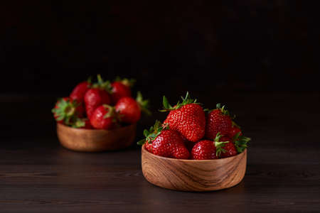 Ripe red strawberries in a round wooden bowl on a wooden table. There is another bowl of strawberries in the background. Brown background, horizontal. Close up, copy space.の写真素材