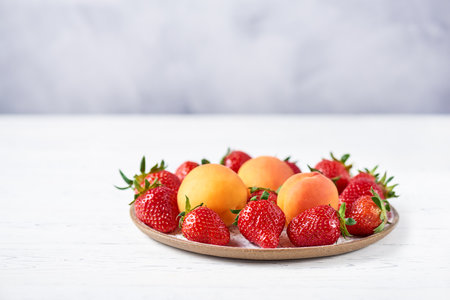 Ripe strawberries and apricots on a flat ceramic plate on a white wooden table. Copy space, close-up.の写真素材