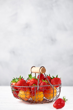 Fresh strawberries and apricots in a metal wicker basket on a white wooden table. gray background. Vertical, copy space, close up.の写真素材