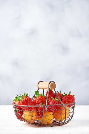 Fresh strawberries and apricots in a metal basket standing on a white table with a gray background. Copy space, close-up.の写真素材
