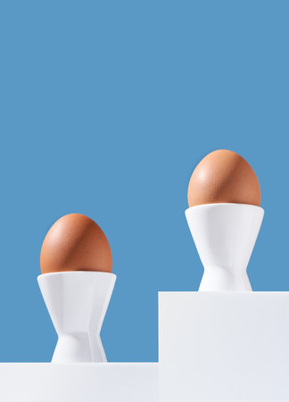 Two brown chicken eggs stand in egg-cups on white catwalks on a blue background. Copy space.の写真素材