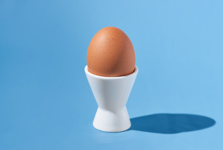 One brown chicken egg stands in a white egg-cup on a blue background. Hard light, copy space, close-up.の写真素材