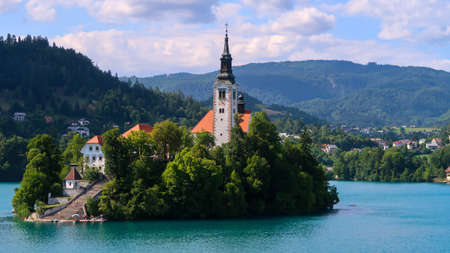 Closeup view of Catholic church situated on an island on Bled lake with mountains and villages on...