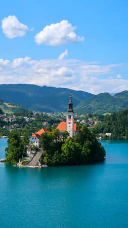 View of Catholic church situated on an island on Bled lake with mountains and villages on the background in the slovenian Alpsの写真素材