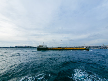 A cargo ship sailing through deep blue waters under a cloudy sky, with a city skyline and historic landmarks in the backgroundの写真素材