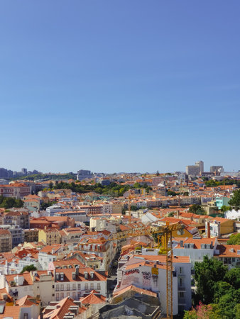A panoramic view of Lisbon, Portugal, featuring red-tiled rooftops, historic buildings, and a construction crane under a clear blue skyの写真素材