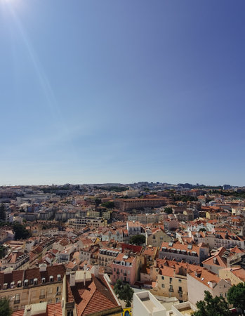 A panoramic view of Lisbon, Portugal, featuring red-tiled rooftops, historic buildings, and a construction crane under a clear blue skyの写真素材
