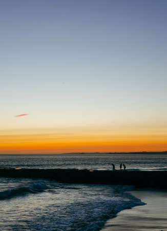 Silhouettes of people walking along the jetty at Praia da Costa da Caparica, Portugal, during a vibrant ocean sunsetの写真素材