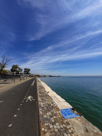 A scenic waterfront pedestrian and bike path in Lisbon, Portugal, alongside the Tagus River under a vibrant blue skyの写真素材