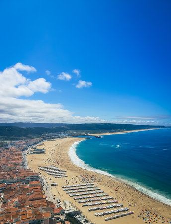 Aerial view of NazarÃ© beach with orange-roofed coastal town, golden sand, turquoise Atlantic waters and umbrellasの写真素材