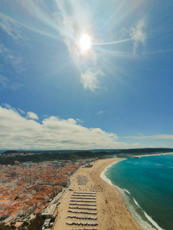 Aerial view of Nazaré beach with orange-roofed coastal town, golden sand, turquoise Atlantic waters and umbrellasの写真素材