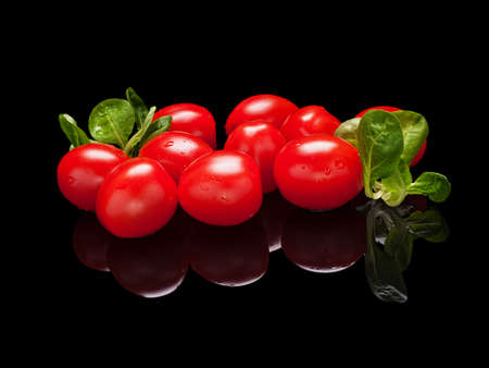 Bunch of small red tomatoes with lettuce leaves isolated on black glossy background with real reflection. Side viewの写真素材