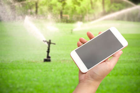 Hand holding smartphone with water sprinkler irrigation of green grass fieldの写真素材