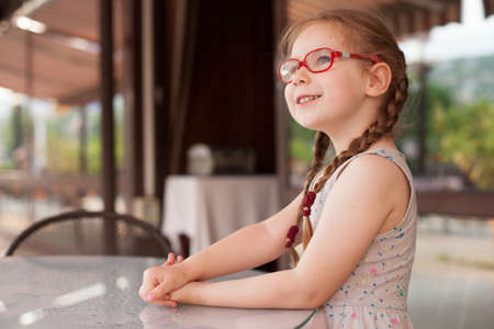 young girl with glasses and dress is sitting at a table in a cafe in the open airの写真素材