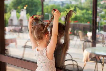 young girl in dress playing with reflection in mirror. laughs picking up pigtails and hairstyles. concept childhood, fun, simplicityの写真素材