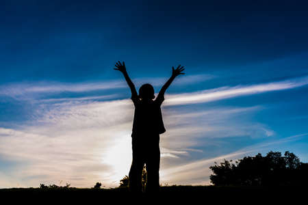 silhouette image of Happy boy raise hand up to the sky on sunset time in background.