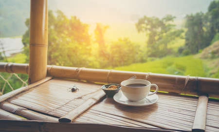 vintage tone image of black coffee in white cup on wooden table to see golden rice field in background.の写真素材