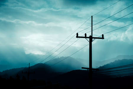 vintage tone image of high voltage electric wire on pole with cloudy sky and mountain before rain on evening time.(focus on background)の写真素材