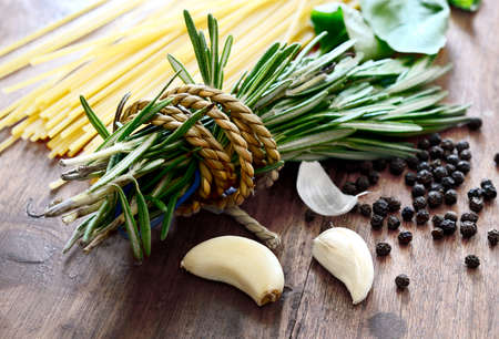 Macro composition of Italian cooking ingredients, garlic, rosemary, basil leaves, spaghetti and black peppercorns on old kitchen table shot in natural light.の写真素材