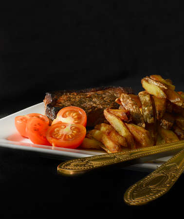 Creatively lit grilled beef steak with French fries and tomatoes against a dark background. Copy...