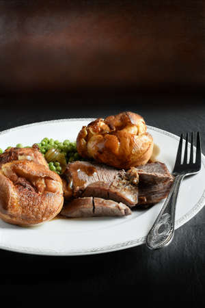 A curated image of topside roast beef, roast potatoes and traditional Yorkshire pudding in a restaurant setting against a dark background with copy space. Concept image for a Sunday lunch menu design.の写真素材