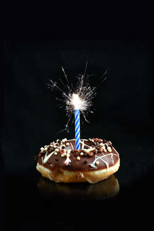 Creative image of a chocolate smothered donut with blue candle sparkler against a black background with copy space. Concept image for celebrations or birthdays.の写真素材