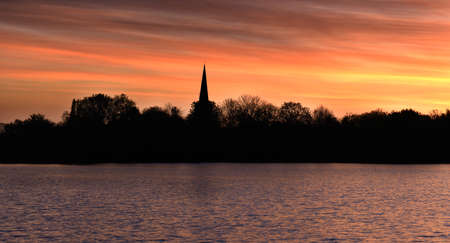 Silhouette of church spire and woodland at dawn sunrise in Nottinghamshire England. Attenborough village church over looking water at the Attenborough Nature Reserve.の写真素材