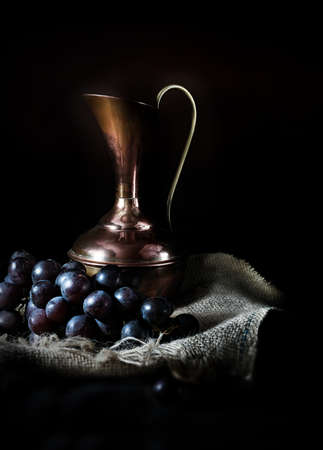 Creatively lit antique jug with black grapes against a dark rustic background with selective focus.の写真素材