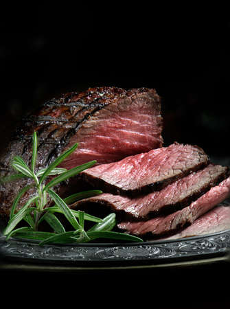 Creatively lit, fresh British roast beef sliced for serving against a dark background with rosemary herb garnish. Generous accommodation for copy space.の写真素材