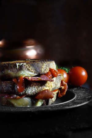 Creatively lit bacon, cheese and tomato stacked sandwich on rye bread with home-made blended tomato relish shot against a dark, rustic background with generous accommodation for copy space.の写真素材