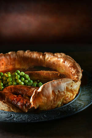 Classic English comfort food, traditional sausages in Yorkshire pudding called toad in the hole. Shot with garden peas and red wine onion gravy against a dark rustic background. Copy space.の写真素材