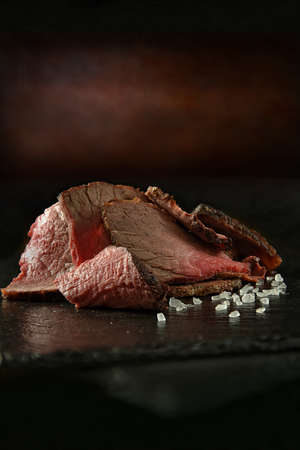 Slices of prime topside roast beef stacked with rock salt on black oiled slate against a rustic, dark background. Generous accommodation for copy space.の写真素材