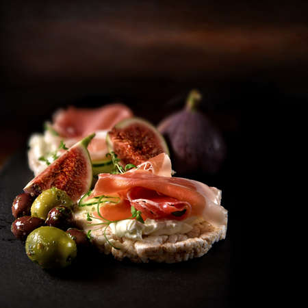 Creatively lit fresh Italian canapes with cream cheese, Serrano cured ham, marinated olives and black figs on organic rice crackers shot against a rustic background with accommodation for copy space.の写真素材