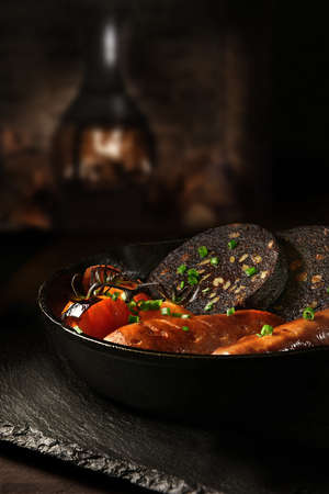Traditional British savoury dish of Black Pudding with sauasages and grilled vine tomatoes in a wrought iron skillet shot against a rustic background with a roaring wood burner. Copy space.の写真素材