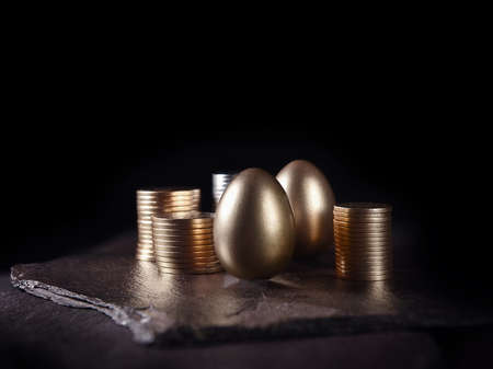 Unique, original creative concept image for financial investments. Gold eggs and stacked coins shot against black, on slate, with copy space.の写真素材