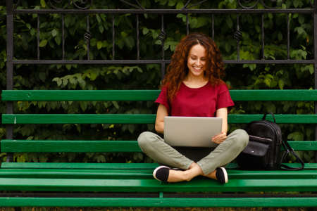 Image of smiling curly young woman sitting on a bench in park and using laptop, isolated outdoor on open air. Horizntal view.の写真素材