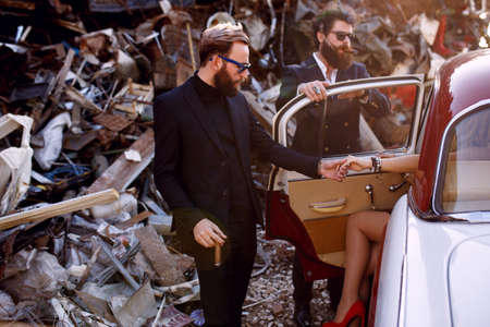 Close up of a stylish brutal bearded mens in black suits and sunglasses smokes cigars near vintage car on the iron dump background and posing at the camera. Gentlemen's club. Horizontal view.の写真素材